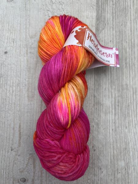 „Handgefärbter Strang in Pink-, Orange- und Gelbtönen, kräftig und warm, fotografiert auf hellem Holzuntergrund mit Herzfasern-Etikett.“